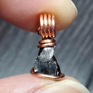 May include: A black, rough-cut gemstone pendant wrapped in copper wire. The pendant is a raw, natural stone with a dark, metallic sheen.