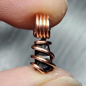 May include: A black crystal pendant wrapped in copper wire. The wire is coiled around the crystal and forms a loop at the top for hanging.