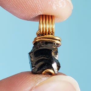 May include: A black, rough, and irregularly shaped stone pendant wrapped in gold wire. The pendant is held by a finger.