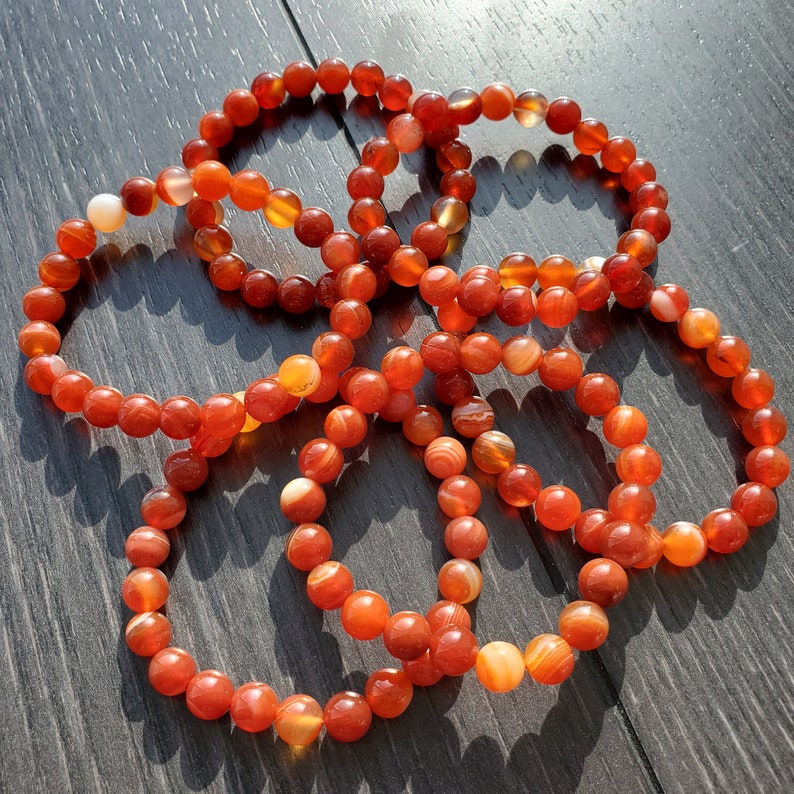 May include: Five carnelian bracelets with orange and red beads. The bracelets are made of natural stone and are stacked on a dark wooden surface.