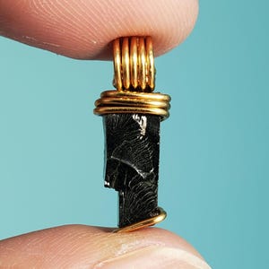 May include: A black, naturally formed,  teardrop-shaped stone pendant wrapped in gold wire. The stone has a unique, striated pattern.