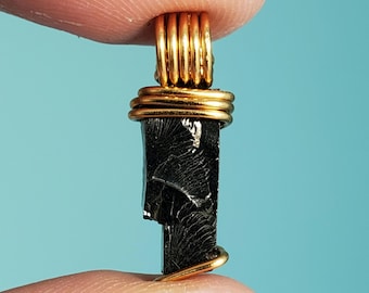Könnte beinhalten: Ein schwarzer, natürlich geformter, tropfenförmiger Steinanhänger, umwickelt mit Golddraht. Der Stein hat ein einzigartiges, gestreiftes Muster.