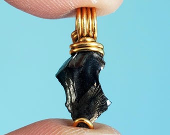 May include: A small, black, irregularly shaped piece of obsidian, wrapped in gold wire, hangs from a gold wire loop. The obsidian has a rough, textured surface.