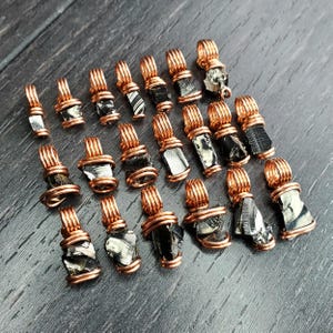May include: A collection of wire-wrapped pendants featuring raw, irregularly shaped black and silver stones. The pendants are crafted with copper-colored wire, showcasing the natural beauty of the stones. The pendants are displayed on a dark gray surface.