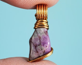 May include: A handcrafted pendant featuring a raw, purple gemstone wrapped in gold-colored wire. The stone has a natural, irregular shape with streaks of white and gray. The pendant is suspended from a gold-colored bail.