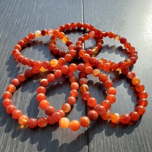 May include: A set of six carnelian bracelets, each made with round, orange-red beads. The bracelets are stacked on top of each other, creating a visually appealing display.