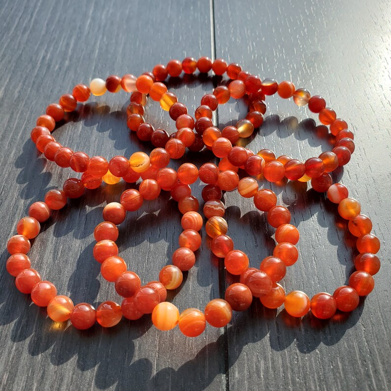 May include: A set of six carnelian bracelets, each made with round, orange-red beads. The bracelets are stacked on top of each other, creating a visually appealing display.