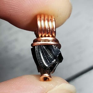 May include: A black obsidian crystal pendant wrapped in copper wire. The pendant is shaped like a rough, natural crystal with a copper loop at the top.