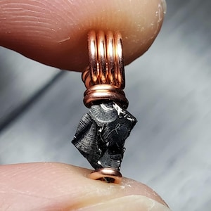 May include: A small, black, irregularly shaped stone pendant with a copper wire wrap. The stone is wrapped with four turns of copper wire.