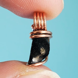May include: A black gemstone wrapped in copper wire, creating a pendant or charm. The gemstone has a rough, natural texture and a few small, gold-colored inclusions.