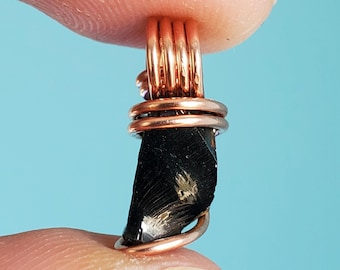 Könnte beinhalten: Ein schwarzer Edelstein, der mit Kupferdraht umwickelt ist und einen Anhänger oder einen Glücksbringer bildet. Der Edelstein hat eine raue, natürliche Textur und einige kleine, goldfarbene Einschlüsse.