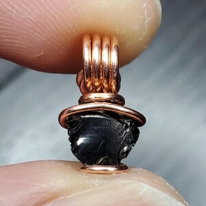 May include: A black gemstone pendant wrapped in copper wire. The pendant is a dark, irregular shape and is suspended from a copper wire loop.