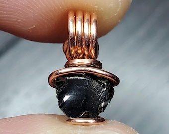 May include: A black gemstone pendant wrapped in copper wire. The pendant is a dark, irregular shape and is suspended from a copper wire loop.