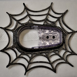 May include: A black and grey spiderweb-shaped decorative frame with a clear plastic insert. The frame has a textured appearance and is designed with a Halloween theme. The clear insert has two circular holes.