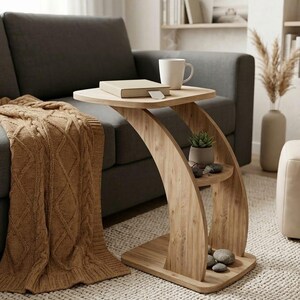 May include: A light-coloured wooden side table with a curved design. The table has a top surface with a book and a mug, and a lower shelf with a small plant and decorative stones. The table is next to a grey sofa and a brown blanket.
