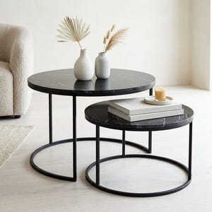 Puede incluir: Una taza de cerámica blanca con un asa negra y el texto "But First, Coffee" en negrita negra. La taza se encuentra sobre un fondo borroso, sugiriendo un ambiente acogedor.