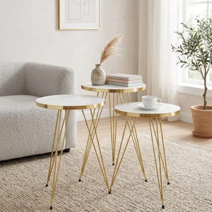 Puede incluir: Un conjunto de tres mesas auxiliares redondas. Las mesas tienen tapas blancas con bordes dorados y patas de metal dorado. Las mesas son de diferentes alturas y tienen un diseño minimalista. Las mesas están en un salón.