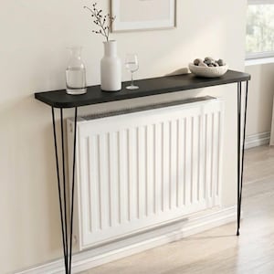 May include: A black console table with hairpin legs, holding a vase, a water carafe, a wine glass, and a bowl of stones. The table is positioned in front of a white radiator. A framed print hangs on the wall above the table.