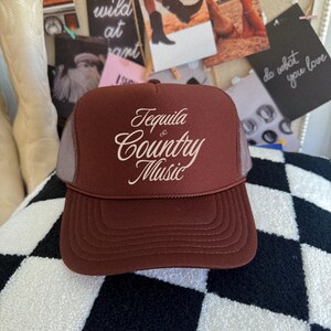 Brown foam front trucker hat with cream “Tequila & Country Music” text, displayed on a patterned surface with western decor.