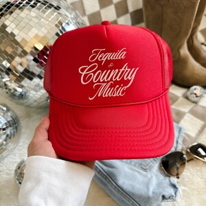 Red foam front trucker hat with cream “Tequila & Country Music” text, shown held in hand with denim and disco ball background.