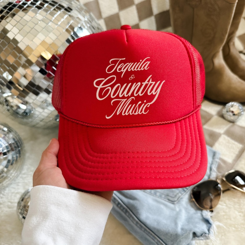 Red foam front trucker hat with cream “Tequila & Country Music” text, shown held in hand with denim and disco ball background.