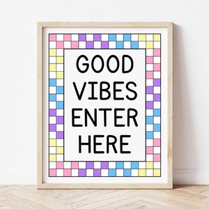 May include: A framed print with the words "GOOD VIBES ENTER HERE" in bold black letters. The print has a colorful checkered border in shades of pink, yellow, blue, and purple. The frame is a light wood color.