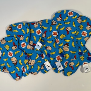 Peut inclure: Quatre coussinets en tissu bleu avec un motif de bananes jaunes et de personnages Donkey Kong. Les coussinets ont des boutons bleus et des étiquettes blanches avec le numéro "4" dessus.