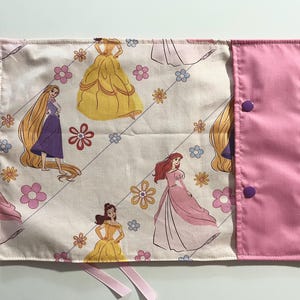 May include: A rectangular fabric roll-up case featuring a cream background with a repeating pattern of cartoon princesses, including Rapunzel, Belle, and Ariel, interspersed with small pink, orange, and blue flowers. It has a solid pink flap with two purple snaps.