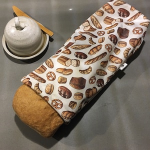 Peut inclure: Un pain brun partiellement enveloppé dans un sac à pain blanc avec un motif de pain brun. Un beurrier en céramique avec un couteau à beurre en bois est à côté du sac à pain.