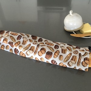 Peut inclure: Un sac à pain long et rectangulaire en tissu avec un motif répétitif de diverses viennoiseries, dont des baguettes, des miches et des bretzels sur fond blanc, doté d'un cordon de serrage noir à une extrémité contenant une baguette.