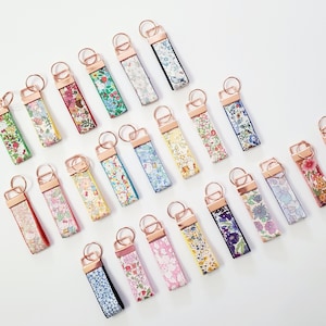 May include: An assortment of floral fabric keychains with rose gold hardware. The keychains feature various colorful floral patterns on a white background. The keychains are rectangular with a loop for keys.