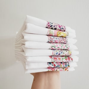 May include: A stack of white linen napkins, each with a floral patterned band. The bands display a variety of colours, including pink, blue, yellow, and green. The napkins are neatly folded and stacked.