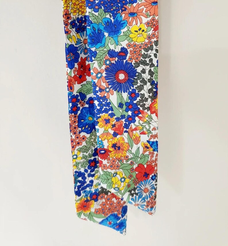 Puede incluir: Bufanda con estampado floral que presenta una vibrante variedad de flores en rojo, azul, amarillo y naranja sobre un fondo blanco. La bufanda est&aacute; hecha de una tela ligera y es ideal para a&ntilde;adir un toque de color a cualquier atuendo.