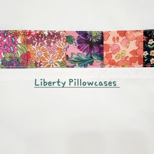 Può includere: Una federa bianca con un bordo in tessuto con motivo floreale colorato. Il bordo presenta un design a patchwork con fiori rosa, arancioni, viola e verdi. Il testo "Liberty Pillowcases" è stampato in verde sulla federa.