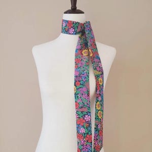 Puede incluir: Una bufanda con estampado floral en tonos rojos, rosas, amarillos y azules est&aacute; drapeada alrededor del cuello de un maniqu&iacute;. El dise&ntilde;o de la bufanda presenta un fondo oscuro con una vibrante variedad de flores. La bufanda es un accesorio largo y delgado.