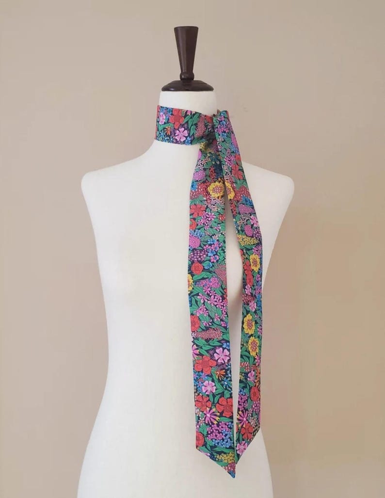 Puede incluir: Una bufanda con estampado floral en tonos rojos, rosas, amarillos y azules est&aacute; drapeada alrededor del cuello de un maniqu&iacute;. El dise&ntilde;o de la bufanda presenta un fondo oscuro con una vibrante variedad de flores. La bufanda es un accesorio largo y delgado.