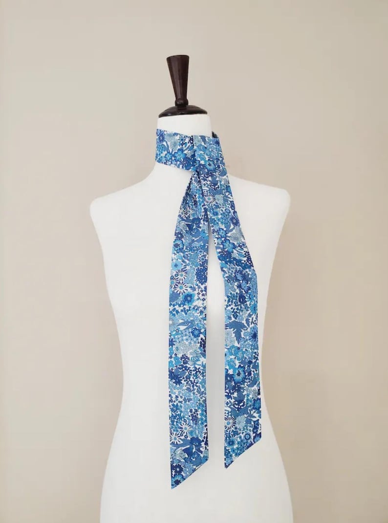 Puede incluir: Una bufanda azul con estampado floral est&aacute; drapeada sobre un maniqu&iacute; blanco. La bufanda presenta una variedad de tonos azules, claros y oscuros, con un delicado dise&ntilde;o floral. La bufanda es de tela suave y es un accesorio elegante.