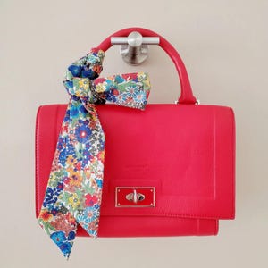 Puede incluir: Un bolso rojo brillante con un asa superior y una bufanda estampada con flores atada. El bolso tiene forma rectangular con cierre de broche. La bufanda presenta un colorido estampado floral.