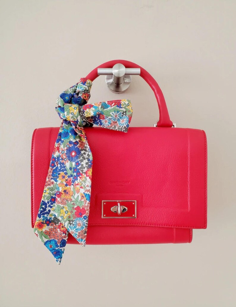 Puede incluir: Un bolso rojo brillante con un asa superior y una bufanda estampada con flores atada. El bolso tiene forma rectangular con cierre de broche. La bufanda presenta un colorido estampado floral.