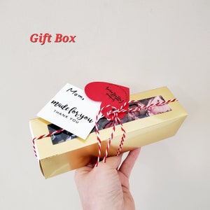 Puede incluir: Una caja de regalo dorada con una ventana transparente, atada con hilo rojo y blanco. La caja contiene art&iacute;culos florales y est&aacute; adornada con una etiqueta en forma de coraz&oacute;n y una etiqueta blanca que dice "Mom, made for you, THANK YOU."