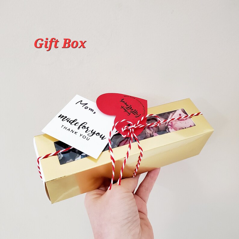 Puede incluir: Una caja de regalo dorada con una ventana transparente, atada con hilo rojo y blanco. La caja contiene art&iacute;culos florales y est&aacute; adornada con una etiqueta en forma de coraz&oacute;n y una etiqueta blanca que dice "Mom, made for you, THANK YOU."