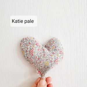 Puede incluir: Un objeto de tela en forma de coraz&oacute;n con un estampado floral en rosa, rojo, amarillo y verde. El texto "Katie pale" est&aacute; en un rect&aacute;ngulo blanco. El objeto est&aacute; sostenido por una mano sobre un fondo blanco.
