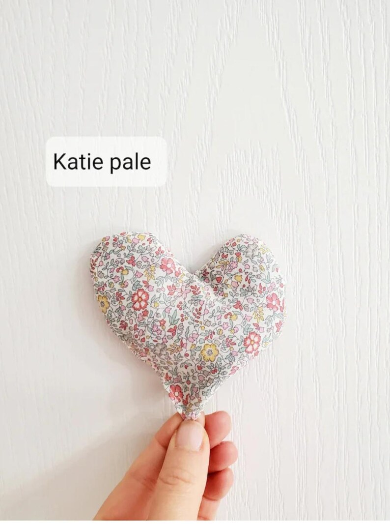 Puede incluir: Un objeto de tela en forma de coraz&oacute;n con un estampado floral en rosa, rojo, amarillo y verde. El texto "Katie pale" est&aacute; en un rect&aacute;ngulo blanco. El objeto est&aacute; sostenido por una mano sobre un fondo blanco.