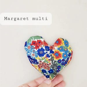 Puede incluir: Un objeto de tela en forma de coraz&oacute;n con un estampado floral en rojo, azul, amarillo y verde. El texto "Margaret multi" se muestra en un rect&aacute;ngulo blanco. El objeto est&aacute; sostenido por una mano.