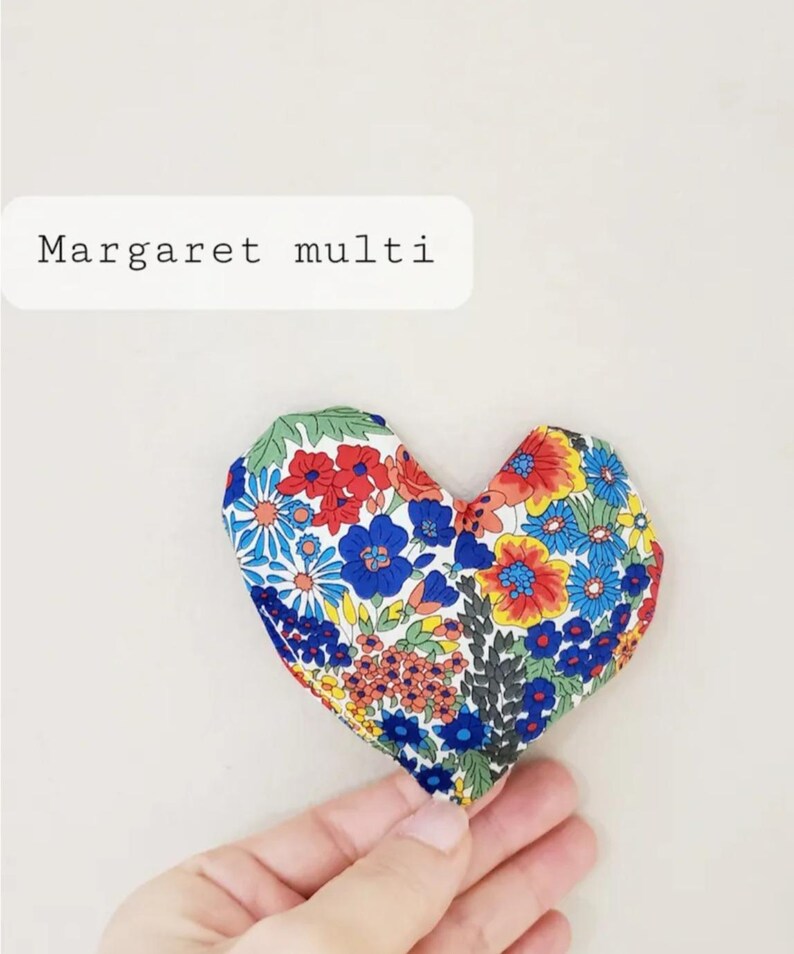 Puede incluir: Un objeto de tela en forma de coraz&oacute;n con un estampado floral en rojo, azul, amarillo y verde. El texto "Margaret multi" se muestra en un rect&aacute;ngulo blanco. El objeto est&aacute; sostenido por una mano.