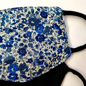 K&ouml;nnte beinhalten: Eine schwarze Gesichtsmaske mit einem blauen und wei&szlig;en Blumendruck. Die Maske hat zwei elastische Ohrschlaufen.