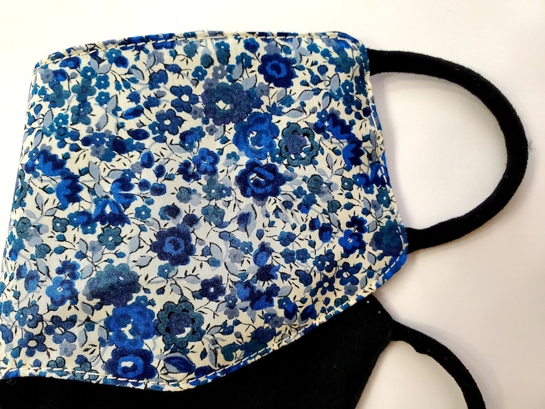 K&ouml;nnte beinhalten: Eine schwarze Gesichtsmaske mit einem blauen und wei&szlig;en Blumendruck. Die Maske hat zwei elastische Ohrschlaufen.