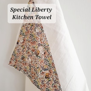 May include: White kitchen towel with a floral and cartoon character patterned accent. The accent features a mix of floral designs and cartoon characters in shades of brown, yellow, and pink. The text "Special Liberty Kitchen Towel" is displayed on the towel.