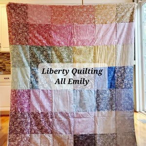 May include: A patchwork quilt made from Liberty fabric with a variety of floral prints in shades of pink, blue, green, and brown. The quilt is hanging on a rod and has the text "Liberty Quilting All Emily" in the center.