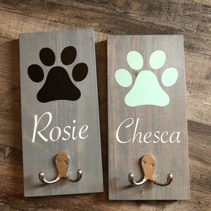 May include: Two wooden dog leash hooks with paw prints and names "Rosie" and "Chesca" in white lettering. The hooks are painted light gray and have silver double hooks.